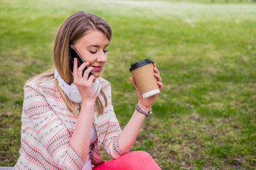 Young woman with coffee to go in hand using a mobile phone and headphones in the park