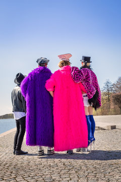 Caucasian men in colorful fake furs and hats standing on a city quay together and holding each other, seen from behind.  