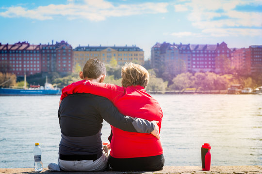 Love couple sitting close together in sunshine on a quay in central Stockholm Sweden, seen from behind. Water and cityscape in the background. 