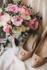 A wedding bouquet of pink peonies, white roses, and eucalyptus, besides beige women's shoes, on which lie the wedding rings. background white cloth