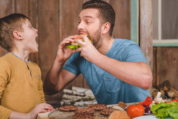 side view of man eating homemade burger with little son near by