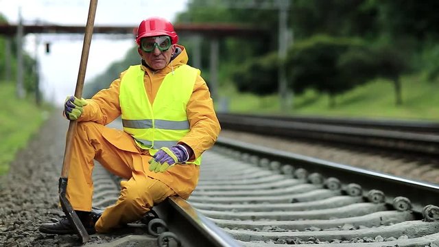 Railwayman in yellow uniform with shovel in hand sits on railway track. Railway construction. Railway worker in red hard hat sits on rail and looks at the camera. Workman with spade on railway track