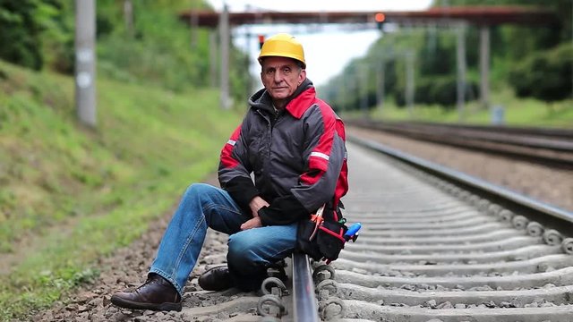 Railwayman in yellow hard hat sits on rail and looks at the camera. Workman on railway track. Railway worker sits on railway line