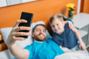 young father and son taking selfie on smartphone at ward, hospital patient care