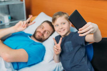 young father and son taking selfie on smartphone at ward, hospital patient care