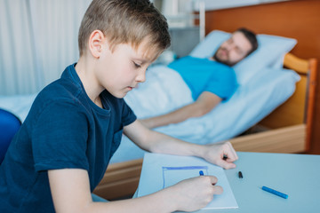 son drawing pictures while sick father laying on hospital bed at ward, dad and son