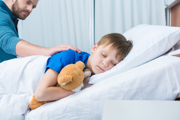 Father touching sick little son lying in hospital bed with teddy bear, dad and son in hospital