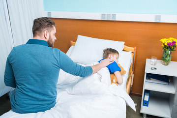Obraz premium High angle view of father touching sick little son lying in hospital bed with teddy bear, dad and son in hospital