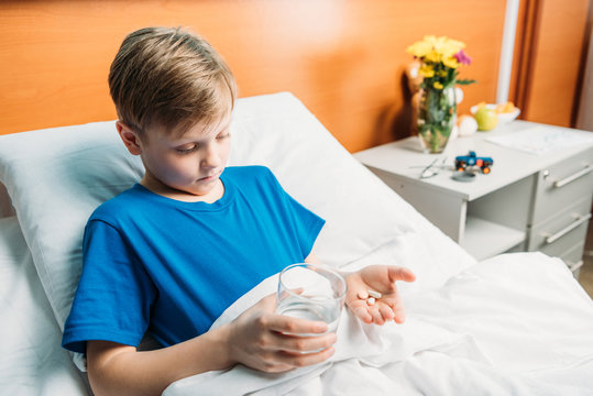 portrait of upset boy holding glass of water and medicines in hospital bed, hospital patient care concept