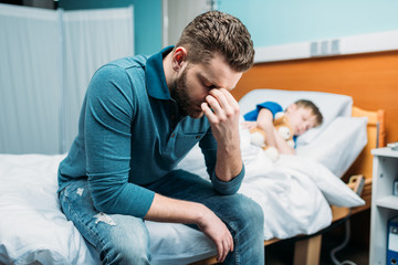 side view of tired dad sitting near sick son in hospital bed