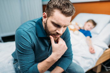 portrait of thoughtful dad sitting near sick son in hospital bed