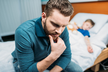 Fototapeta premium portrait of thoughtful dad sitting near sick son in hospital bed