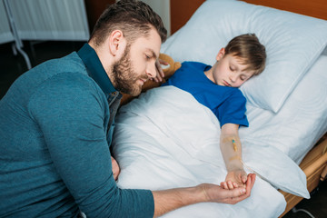 portrait of pensive dad sitting near sick son in hospital bed