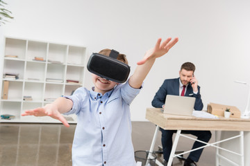 Boy using virtual reality glasses while his father businessman working in office