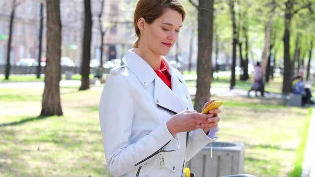 SMS. Young blonde woman reads messages on cellphone sitting on bench in park