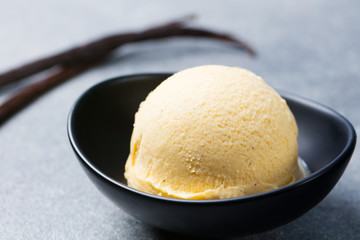 Vanilla, caramel ice cream in black bowl with vanilla pods. Grey stone background.