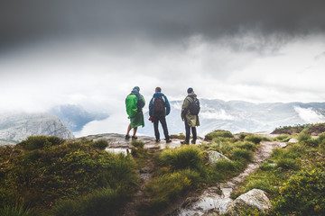 Obraz premium Three male hikers staying at peak summit, looking away from above at beautiful landscape over Preikestolen, famous and very popular touristic landmark in Norway, Scandinavia. Bad weather conditions.
