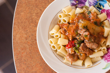 Close up of american chop suey - pasta dish with beef, tomato sauce, mozarella cheese, spices and parsley. Italian-American cuisine. White plate with steaming hot pasta and pan with rest dish on back