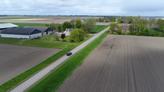 Aerial flight over polder landscape following car on rural road driving fast over the small paved road furthermore showing brown plowed farm fields left and right and also small farm in middle 4k