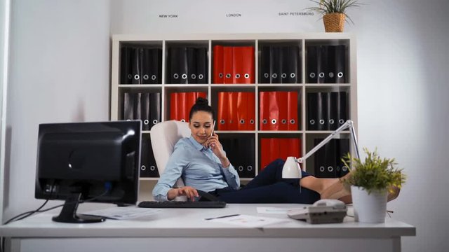 Satisfied relaxed businesswoman sitting in a chair with legs over the table and hands in the head enjoying her new job at office. Iphone ipad table pc monitor phone. Glidetrack dolly teal orange shot