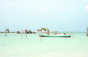 Fototapeta na wymiar Empty fishing boat docked at the turquoise colored ocean coast near white sand beach in the afternoon with rocks on the ocean horizon in Belitung, Indonesia