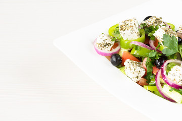 Greek salad with fresh tomato, cucumber, red onion, basil, feta cheese, black olives, Italian herbs and olive oil in white dish on white wooden table.
