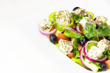 Greek salad with fresh tomato, cucumber, red onion, basil, feta cheese, black olives, Italian herbs and olive oil in white dish on white wooden table.