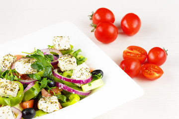 Greek salad with fresh tomato, cucumber, red onion, basil, feta cheese, black olives, Italian herbs and olive oil in white dish on white wooden table.
