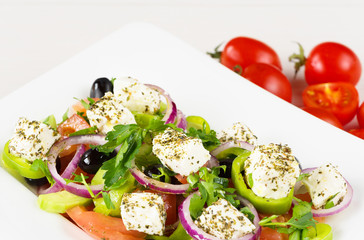 Greek salad with fresh tomato, cucumber, red onion, basil, feta cheese, black olives, Italian herbs and olive oil in white dish on white wooden table.