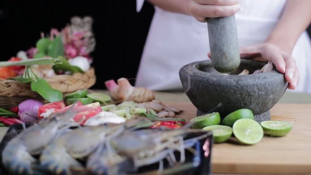 Female hand pulverizing Thai herb in the mortar to make Tom Yum Kung with all spices and prawns beside on the chopping board.