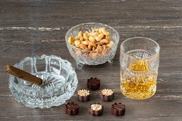 Glass of whiskey, a cuban cigar, nuts and chocolates on a grey wooden table. Close-up view.