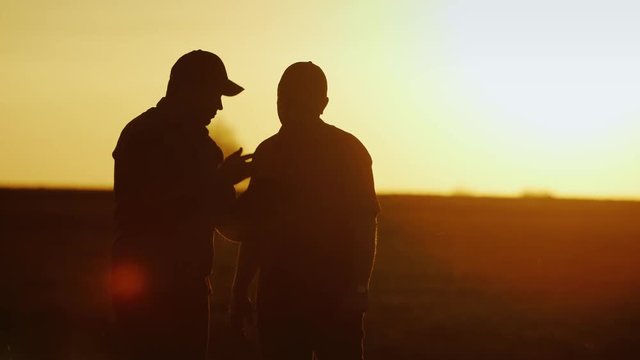 Deal in agribusiness. Two male farmer communicate on the field, use a tablet - shake hands. Silhouettes at sunset