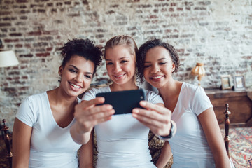 Pretty girls having fun and make selfie sitting near the bed.