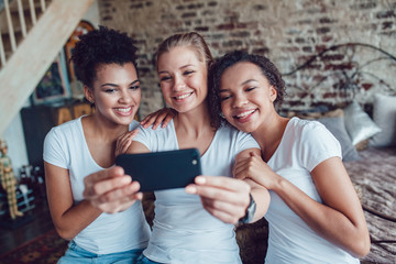 Pretty girls having fun and make selfie sitting near the bed. Two afro-americans and one european girl.