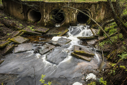 River dam made of pipes under a bridge in a forest road with running water.