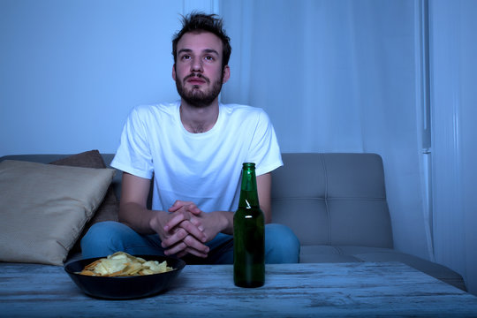 Young man watching a sad movie at night with chips and beer.
