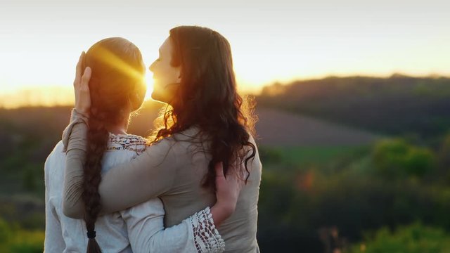 Mom gently kisses her daughter's temple. Together they admire the beautiful sunset. Back view