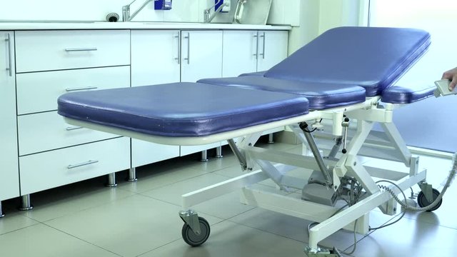 Blue couch for patients stand in premise in clinic. Young man dressed in white uniform raises folding medical squab with brilliant leather coating on wheels with automatic device holding miniature