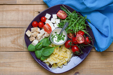 Big portion of farfalle pasta salad with cherry tomatoes, baby spinach leaves, chicken meat, feta cheese, rocket leaves and white sauce served on wooden table with blue napkin