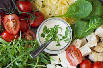 White mayonnaise sauce served with fresh rocket salad leaves, red cherry tomatoes, farfalle pasta, baby spinach leaves, feta cheese cubes and chicken meat. Top view. Salad dressing