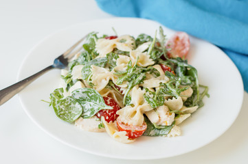 Portion of pasta salad with rucola leave, baby spinach, farfalle, sliced cherry tomatoes and chicken meat dressed with white sauce and served on white plate with fork and blue napkin
