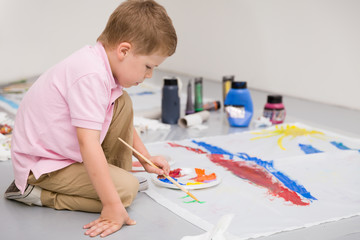 Cute kid boy sitting on the floor and drawing with colorful paints. Child making flag craft. Education, creativity, school activities.