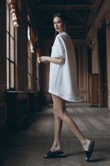 Beautiful sexy model in short white dress and slippers in step pose in theatre corridor. Brunette girl with long slim legs walking in classic museum interior. Elf hairstyle. Fashion photo backside