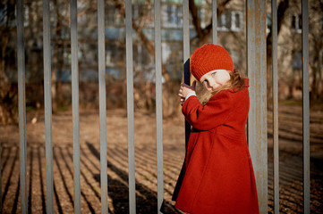 Baby 3 years with long hair. In a red beret and coat stands on the street in the sunshine, near the fence