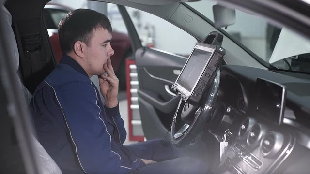 Automaster sitting in a car and thinking how to do chip tuning. the service center, mechanic in blue uniform is in the driver's seat in the car and directly looks at the diagnostic device that is