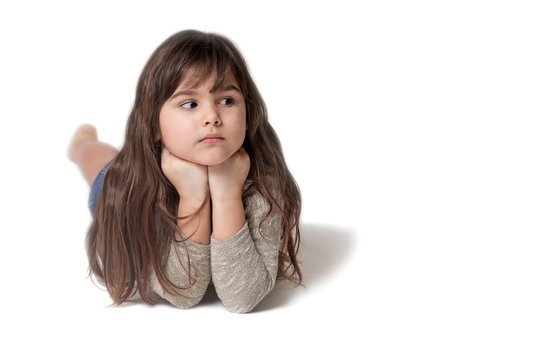 Brunette long haired little girl is lying on the gray background. The girl is supporting  her chin with both hands and is looking at the empty place of the photo ready for your text. 