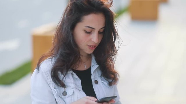 Brown-haired beatiful caucasian girl in elegant light white jeans coat with pretty smile communicating via smartphone phablet Steadicam slow motion video