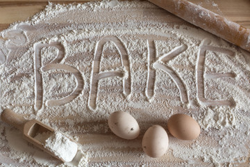 Bakery background. Flour artwork with food eggs, wooden spoon with flour and wooden rolling pin, fun background with the word Bake, and in scattered flour on a wooden tabletop