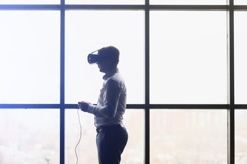 The VR headset design is generic and no logos, Man wearing virtual reality goggles watching movies or playing video games. Standing near the window, side view, background,,flares effect