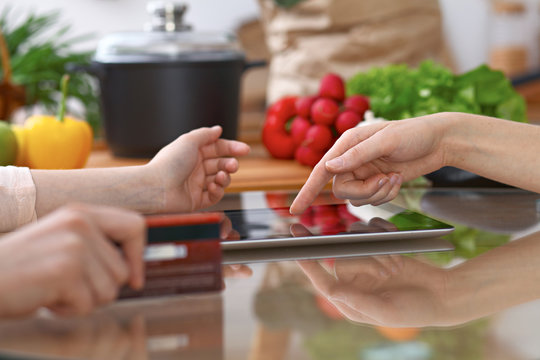 Human hands of two female persons using touchpad in the kitchen. Closeup of two women are making online shopping by tablet computer and credit card. Cooking and shopping concept